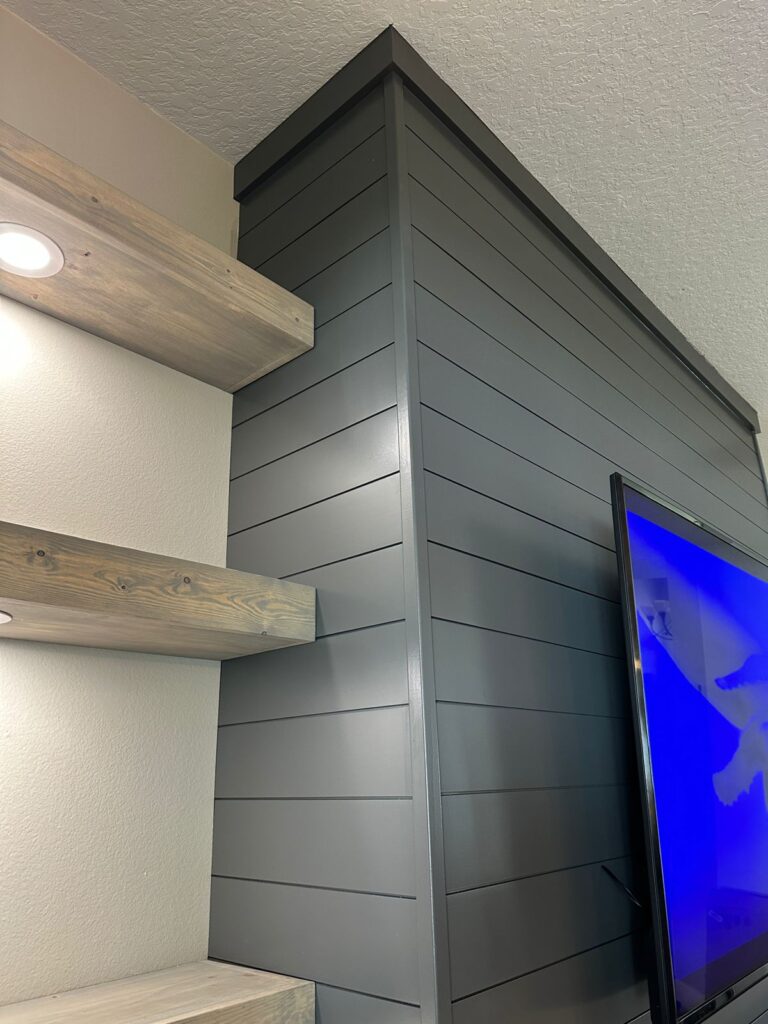 Modern black shiplap TV wall with floating wood shelves and built-in media feature detail in a contemporary living room. Custom accent wall installation in Orlando, Florida.