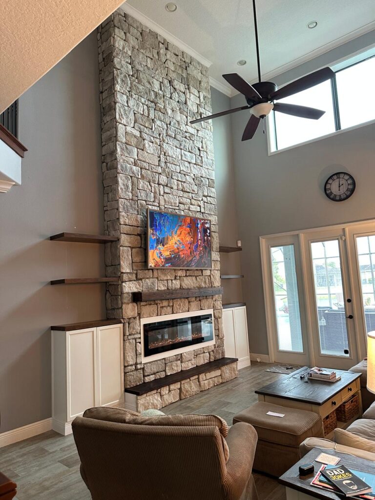 Custom tall stone fireplace wall with floating shelves, electric fireplace and mounted TV in luxury Florida living room