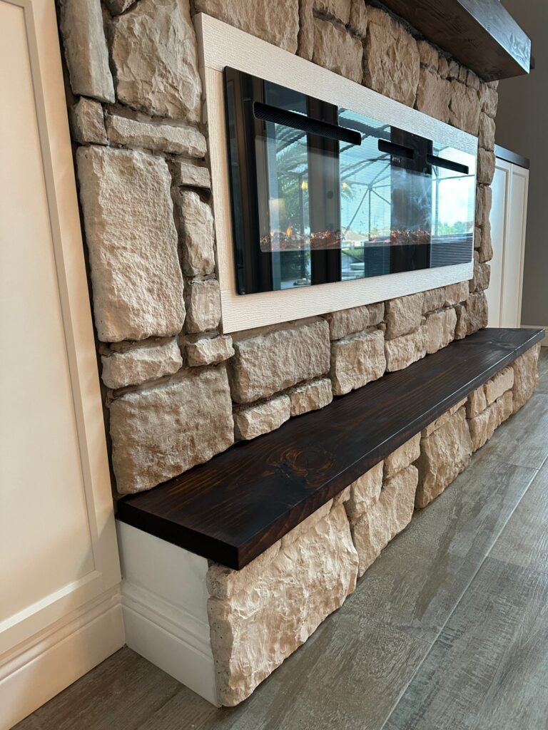 Close-up of custom stone fireplace wall with dark floating wood mantel and modern electric fireplace in Florida home